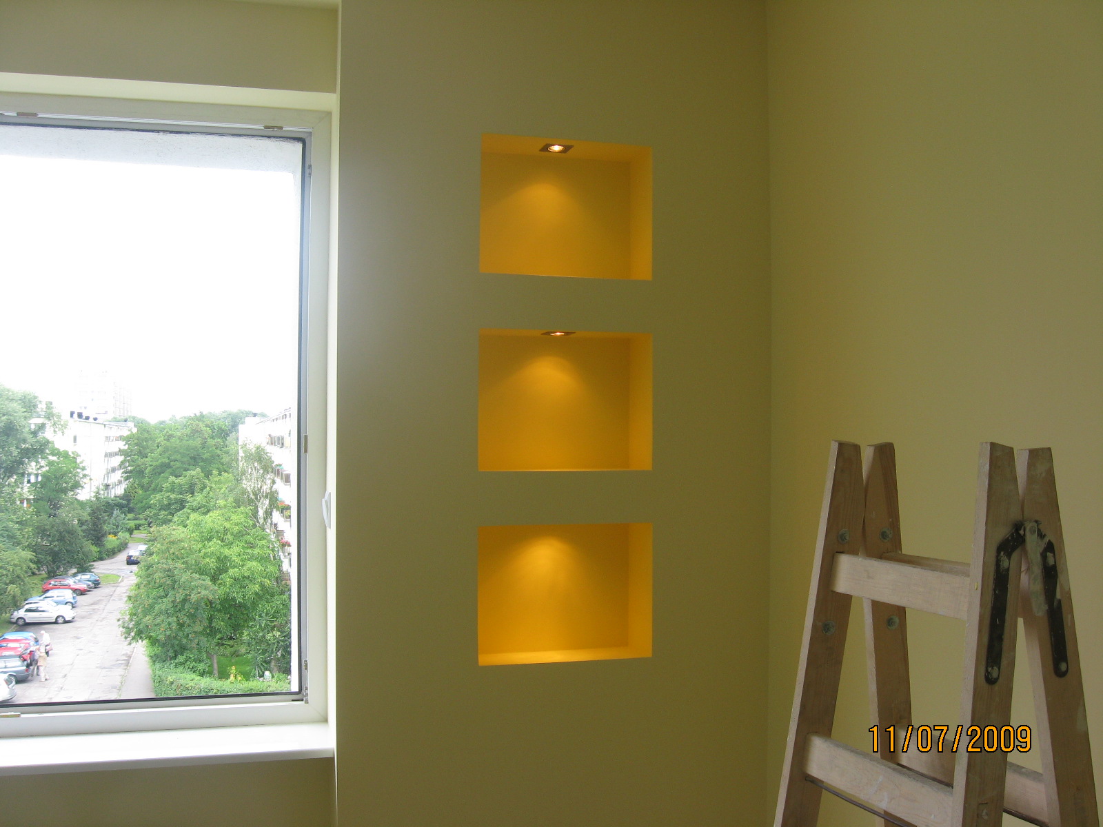 Ściana z trzema podświetlonymi niszami, widok z okna na drzewa i budynki, w rogu drabina malarska z datą 11/07/2009.