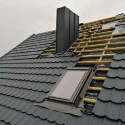 Montaż okna dachowego w szarym dachu z metalowej dachówki, widoczna konstrukcja drewniana i komin w tle, pochmurne niebo.