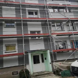 Wielopiętrowy budynek mieszkalny w trakcie prac elewacyjnych, osłonięty rusztowaniami z widocznymi fragmentami pomalowanej w pasy elewacji. Okna zabezpieczone folią, wejście osłonięte plandeką,...