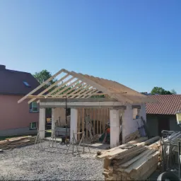 Wznoszenie ścian garażu z bloczków betonowych z widoczną konstrukcją więźby dachowej, materiały budowlane składowane na placu budowy, w tle dom mieszkalny.