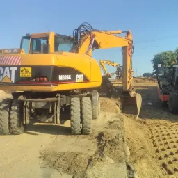 Żółta koparka kołowa CAT M316C w trakcie prac ziemnych, w tle widoczny walec HAMM i druga koparka, teren wyrównany z charakterystycznym wzorem.