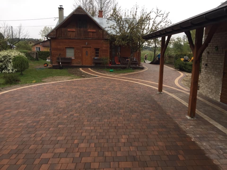 Ułożona kostka brukowa w odcieniach brązu tworząca krętą ścieżkę przed drewnianym domem z gankiem i zadaszeniem, jasne obrzeża kostki podkreślają kształt alejki.