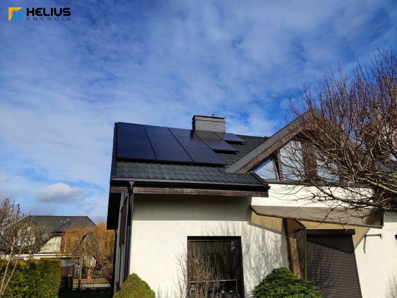 Czarny dach z panelami fotowoltaicznymi na białym domu jednorodzinnym pod błękitnym, lekko zachmurzonym niebem. Widoczny logo firmy Helius Energia w lewym górnym rogu.
