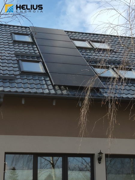 Czarny panel słoneczny na dachu domu z dachówką, widoczne okna dachowe i logo firmy HeliuS Energia w lewym górnym rogu.
