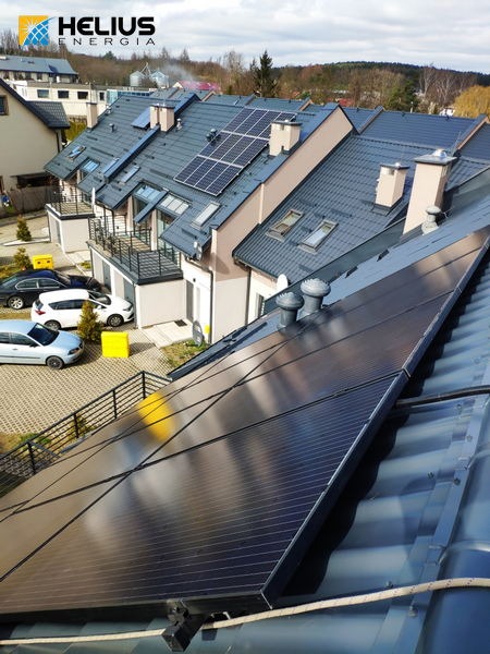 Część dachu z panelami słonecznymi na tle szeregu domów w zabudowie bliźniaczej, widoczny znak firmowy na górze kadru.