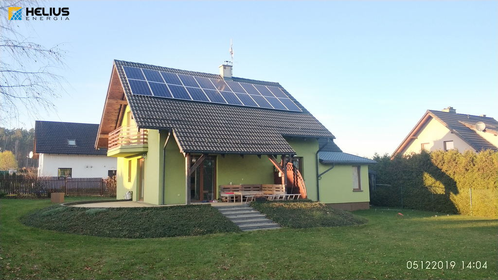 Dom jednorodzinny z panelami słonecznymi na dachu, widoczny z poziomu trawnika w słoneczny dzień. Na dachu antena satelitarna.