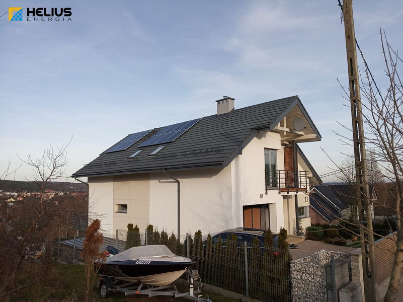 Nowoczesny dom jednorodzinny z ciemną dachówką i panelami słonecznymi, widok z zewnątrz, na pierwszym planie łódź motorowa na przyczepie.