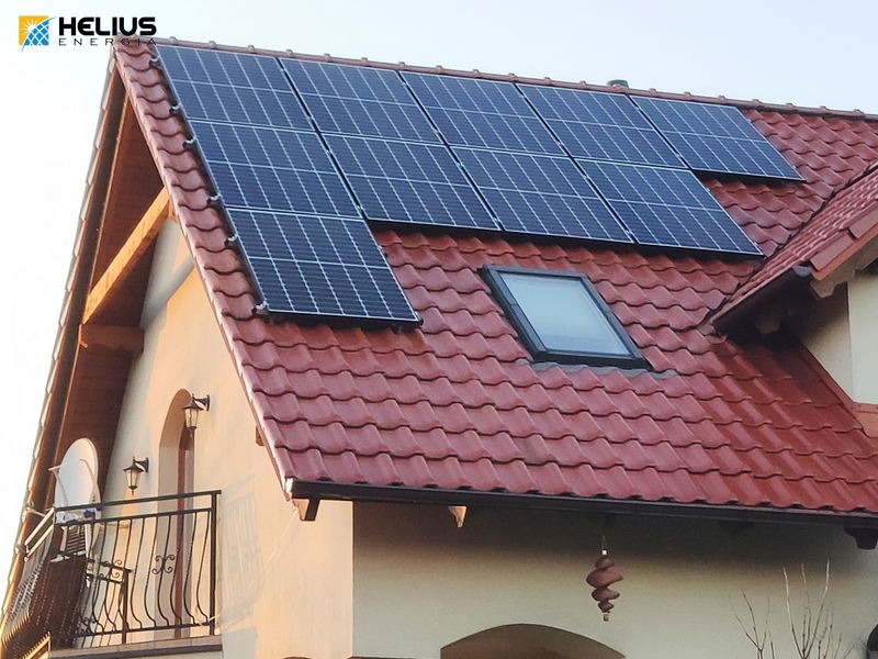 Panele słoneczne na czerwonym dachu domu jednorodzinnego z oknem dachowym; logo firmy Helius Energia w lewym górnym rogu.