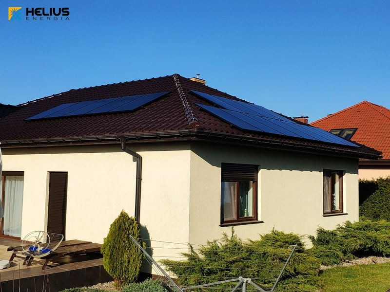 Dom jednorodzinny z brązowym dachem, na którym zamontowano panele słoneczne. Widoczny ogród z krzewami i drzewkami iglastymi. Na dachu dwa zestawy paneli ułożone w rzędach.