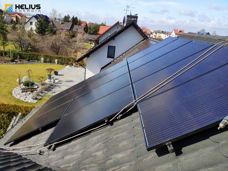 Panele słoneczne na dachu domu jednorodzinnego, widoczne kable instalacyjne, w tle zabudowa mieszkalna i trawnik.