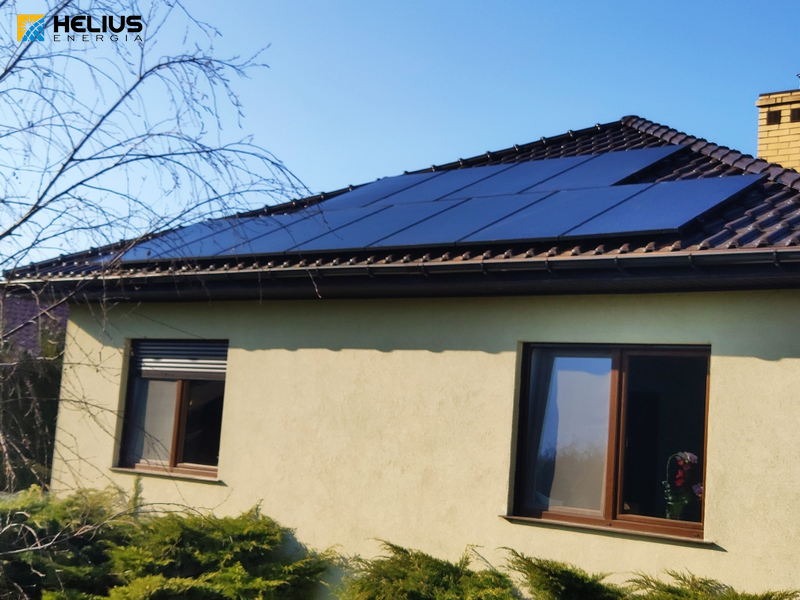 Panele słoneczne na brązowym dachu domu jednorodzinnego z beżową elewacją, widoczne krzewy i błękitne niebo.