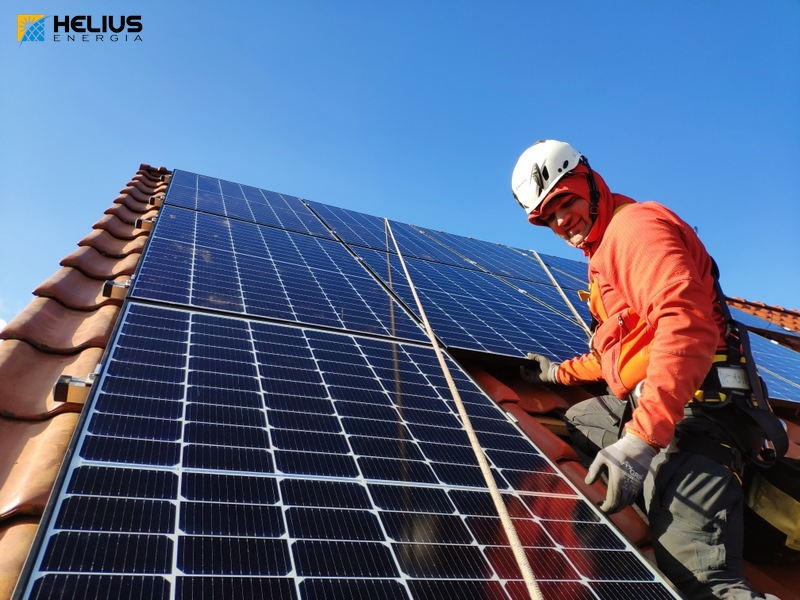 Instalator w kasku montuje panele słoneczne na brązowym, spadzistym dachu, widok z dołu na tle błękitnego nieba.