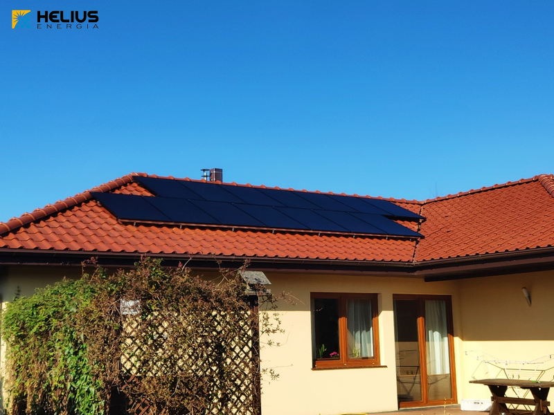 Czerwony dach z dachówki ceramicznej z instalacją paneli słonecznych, widok z poziomu ogrodu, błękitne niebo w tle, logo firmy w lewym górnym rogu.