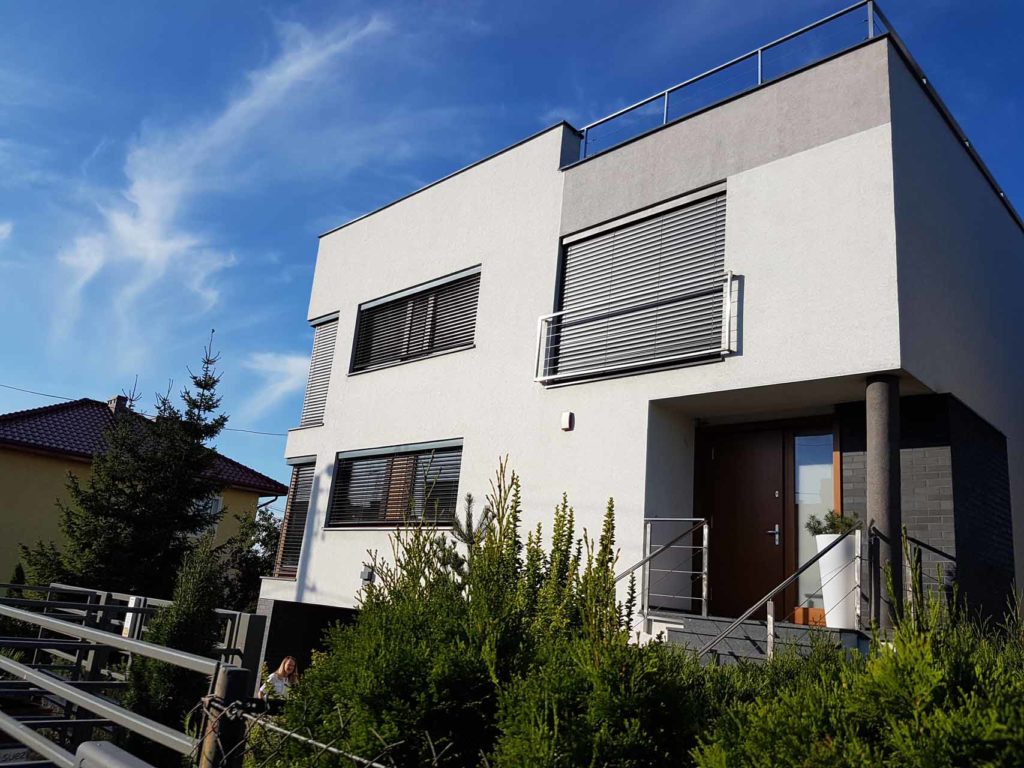 Nowoczesny dom z szarymi roletami zewnętrznymi, białą elewacją i antracytowymi elementami wykończeniowymi, widziany z poziomu ogrodu w słoneczny dzień.