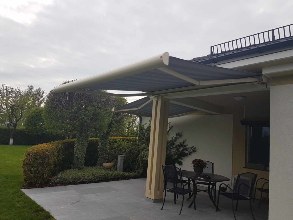 Zewnętrzny taras z rozkładaną markizą w kolorze szarym, wspartą na beżowych kolumnach, z czarnym zestawem mebli ogrodowych i widokiem na zadbany trawnik z krzewami.