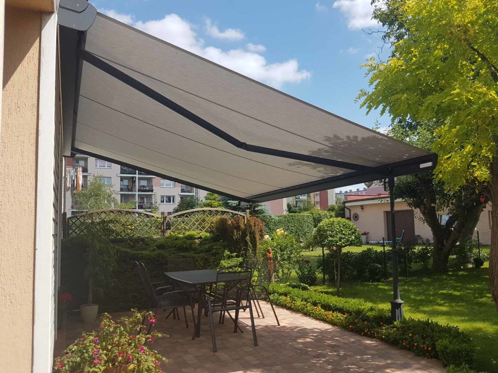 Zewnętrzna, rozkładana markiza tarasowa z czarną ramą i szarym materiałem, zacieniająca patio z ceglaną podłogą, stolikiem i krzesłami, otoczona zielenią ogrodu i budynkami w tle.