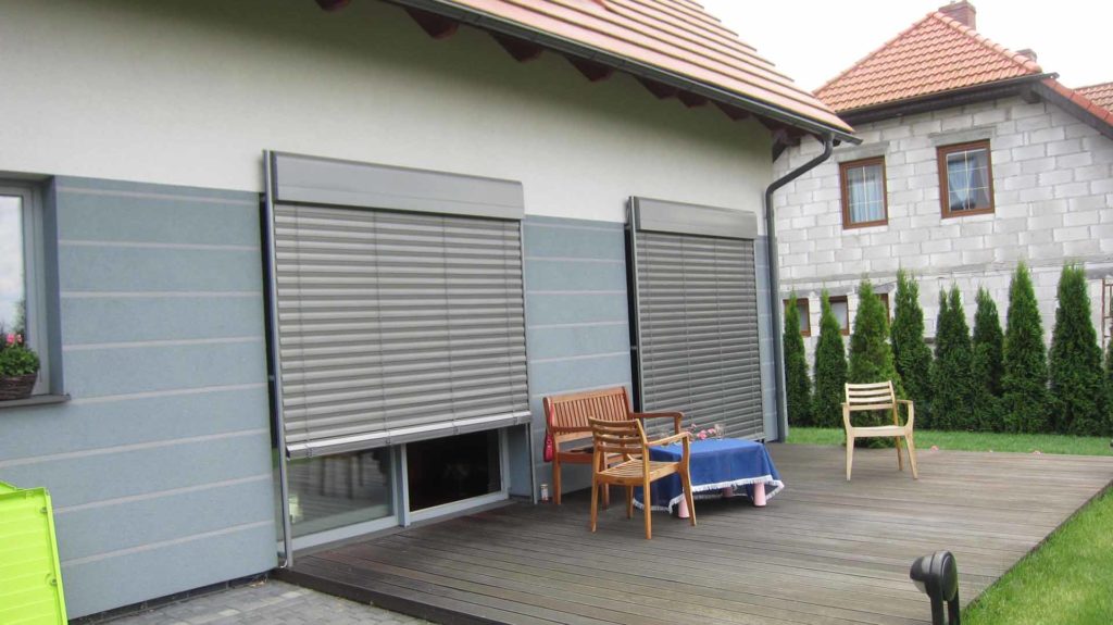 Zewnętrzne rolety okienne w kolorze szarym, częściowo opuszczone, na tle jasnego domu z drewnianym tarasem i meblami ogrodowymi.
