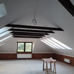 Poddasze w trakcie wykańczania: białe ściany i sufit, drewniane belki stropowe, okna dachowe wpuszczające światło, fragment odsłoniętej cegły na ścianie, widoczne przygotowania do dalszych prac.