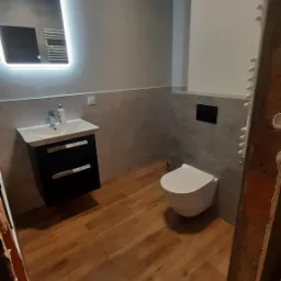 Nowoczesna łazienka z drewnopodobną podłogą, podświetlanym lustrem nad czarną szafką z umywalką i wiszącą toaletą, wykończona szarymi płytkami.