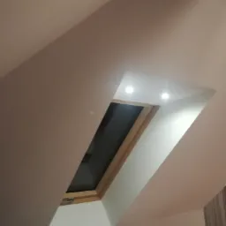 Poddasze z oknem dachowym w drewnianej ramie, widoczne oświetlenie punktowe w suficie i fragment grzejnika.