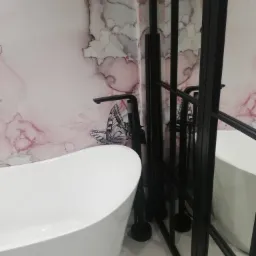 Nowoczesna łazienka z wolnostojącą białą wanną, czarną armaturą i ścianą z motywem marmuru oraz naklejką motyla, oddzielona czarną, przeszkloną ścianką działową.