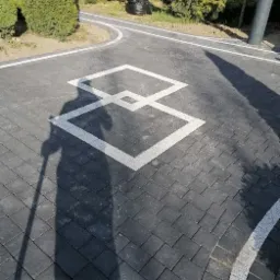 Ułożona kostka brukowa w kolorze grafitowym z białym wzorem geometrycznym, cień osoby z kijem, widoczne elementy otoczenia: trawnik, drzewa, ławka, ścieżka.