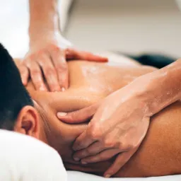 Olejowany tors mężczyzny podczas manualnego rozluźniania mięśni szyi i pleców przez terapeutę. Dłonie terapeuty w trakcie masażu, widoczne z bliska.