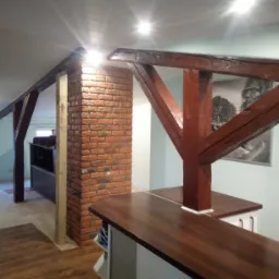 Wykończenie poddasza z drewnianymi belkami, imitacją cegły na filarze i brązowym blatem, oświetlenie punktowe w suficie.