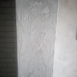 Teksturowana, biała ściana z efektem dekoracyjnym, sąsiadująca z fragmentem ściany wyłożonej cegłą w odcieniach szarości i gładką ścianą w tle.