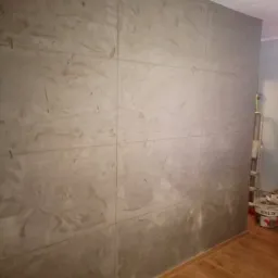 Ściana pokryta szarą, imitującą beton, płytą. Widoczne łączenia płyt, drewniana podłoga i drabina w tle.