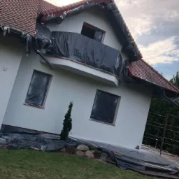 Dom jednorodzinny w trakcie malowania elewacji, zabezpieczony folią, widoczne okna i balkon również osłonięte, częściowo pomalowana ściana na jasny kolor.