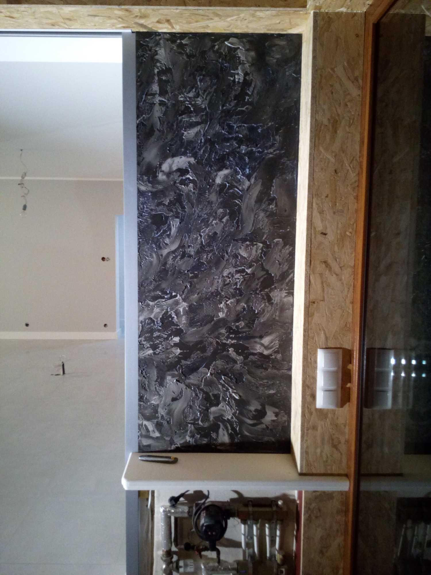 Ściana z dekoracyjną szpachlą w odcieniach szarości, widoczny fragment instalacji grzewczej i surowa płyta OSB