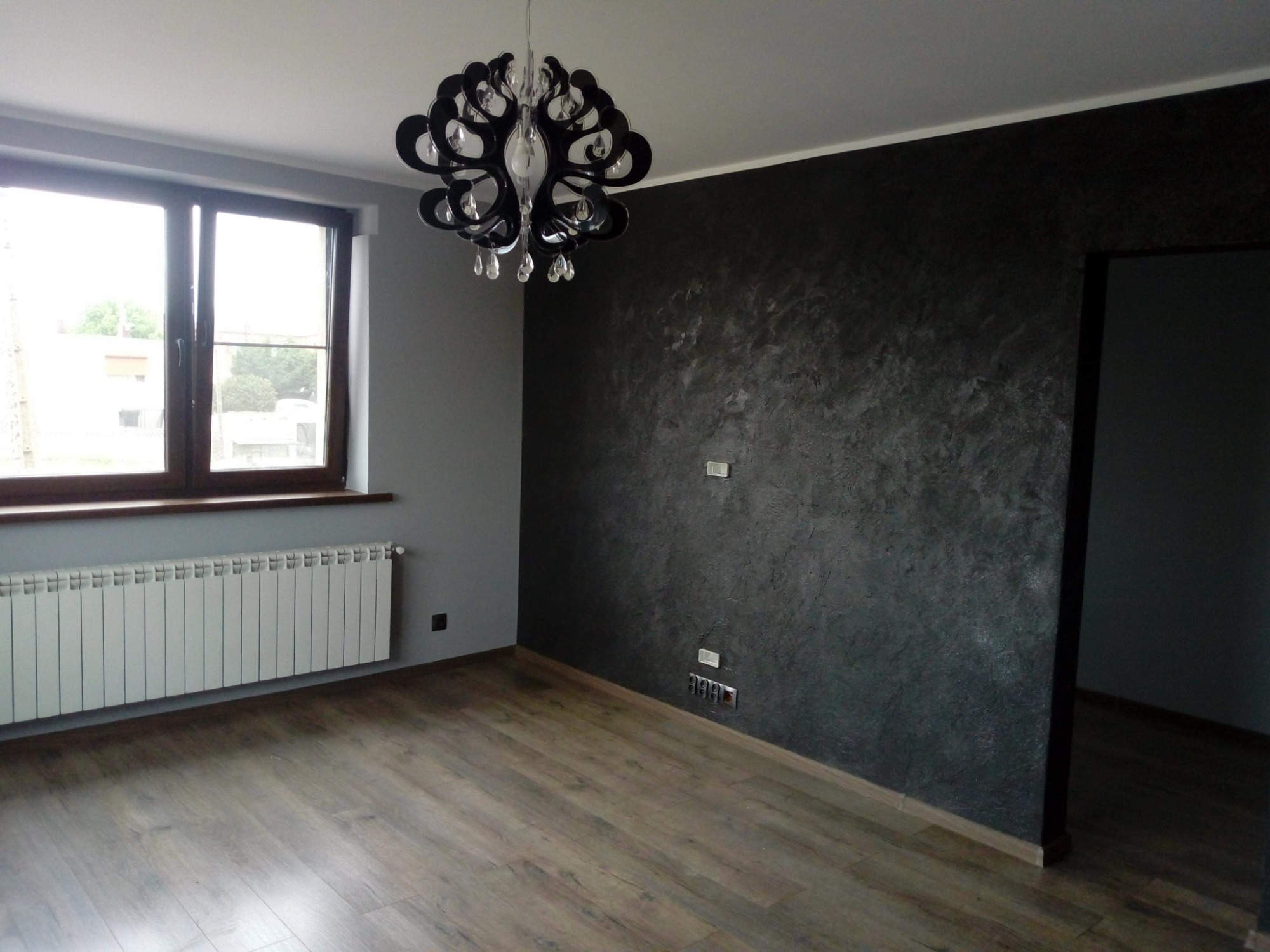 Pokój po remoncie: szara ściana z oknem, grzejnik, ciemna ściana z efektem dekoracyjnym, ozdobny żyrandol i drewniana podłoga.