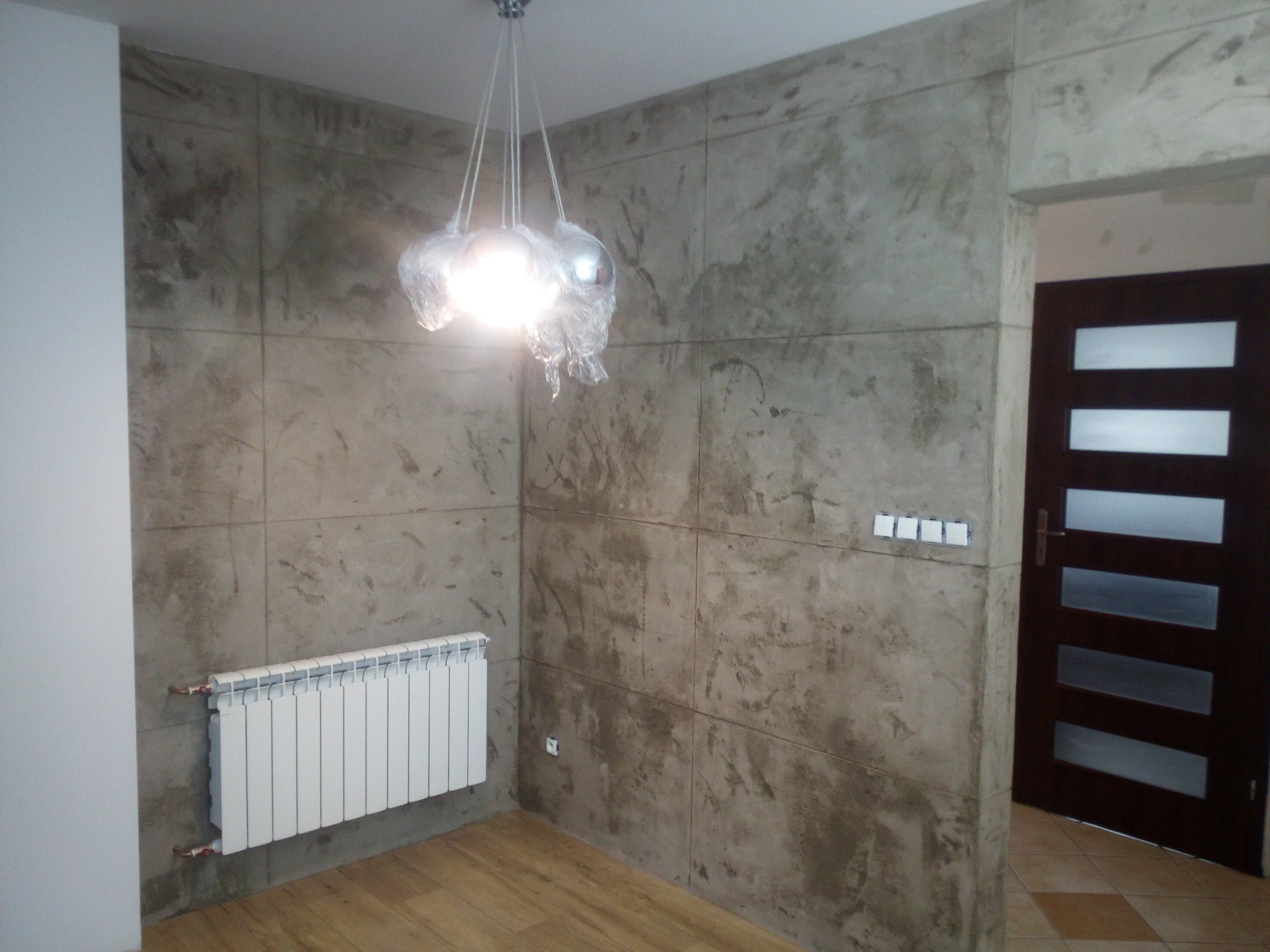 Pokój z dekoracyjną ścianą w odcieniach szarości, podzieloną na kwadraty, z białym grzejnikiem i lampą wiszącą jeszcze w folii ochronnej, widoczna drewniana podłoga i drzwi z szybami.