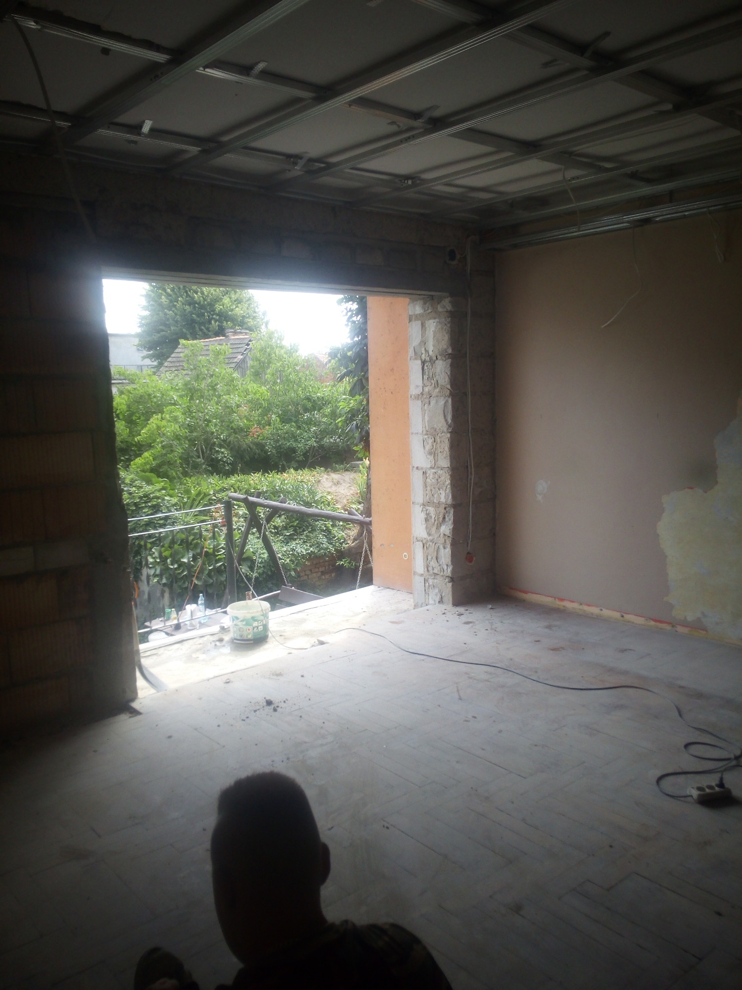 Wnętrze w trakcie prac wykończeniowych: surowa ściana, częściowo położona gładź, widoczna konstrukcja sufitu podwieszanego, okno z widokiem na zieleń.