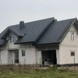 Surowy stan domu jednorodzinnego w trakcie budowy, widoczny czarny dach z dachówki, białe ściany z pustaków, częściowo zamontowane okna, teren wokół budynku niezagospodarowany, jesienna sceneria.