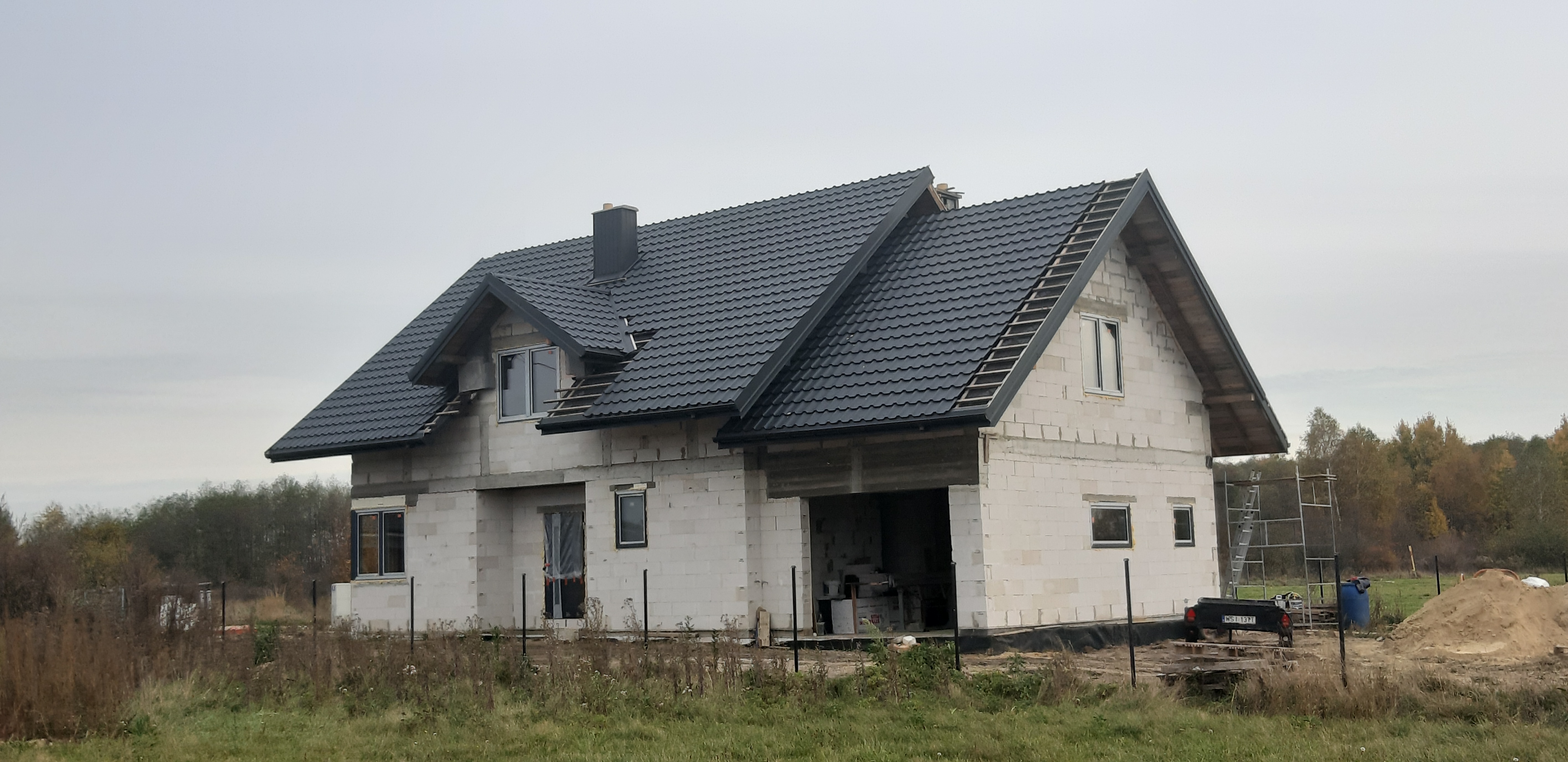 Surowy stan domu jednorodzinnego w trakcie budowy, widoczny czarny dach z dachówki, białe ściany z pustaków, częściowo zamontowane okna, teren wokół budynku niezagospodarowany, jesienna sceneria.