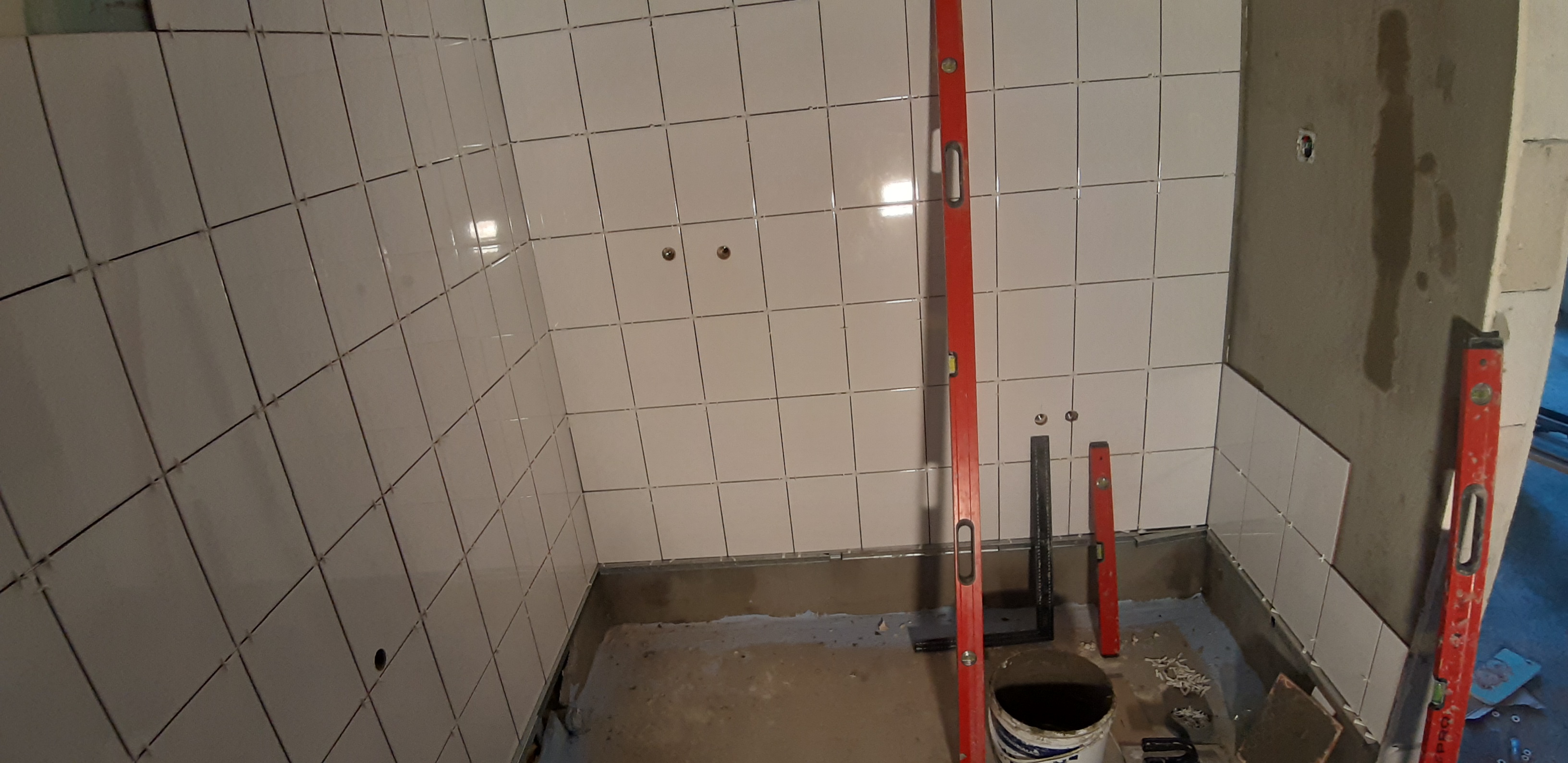 Świeżo ułożone, białe, kwadratowe płytki na ścianach w rogu pomieszczenia, z widocznymi poziomnicami i narzędziami, ukazujące etap prac wykończeniowych w łazience.