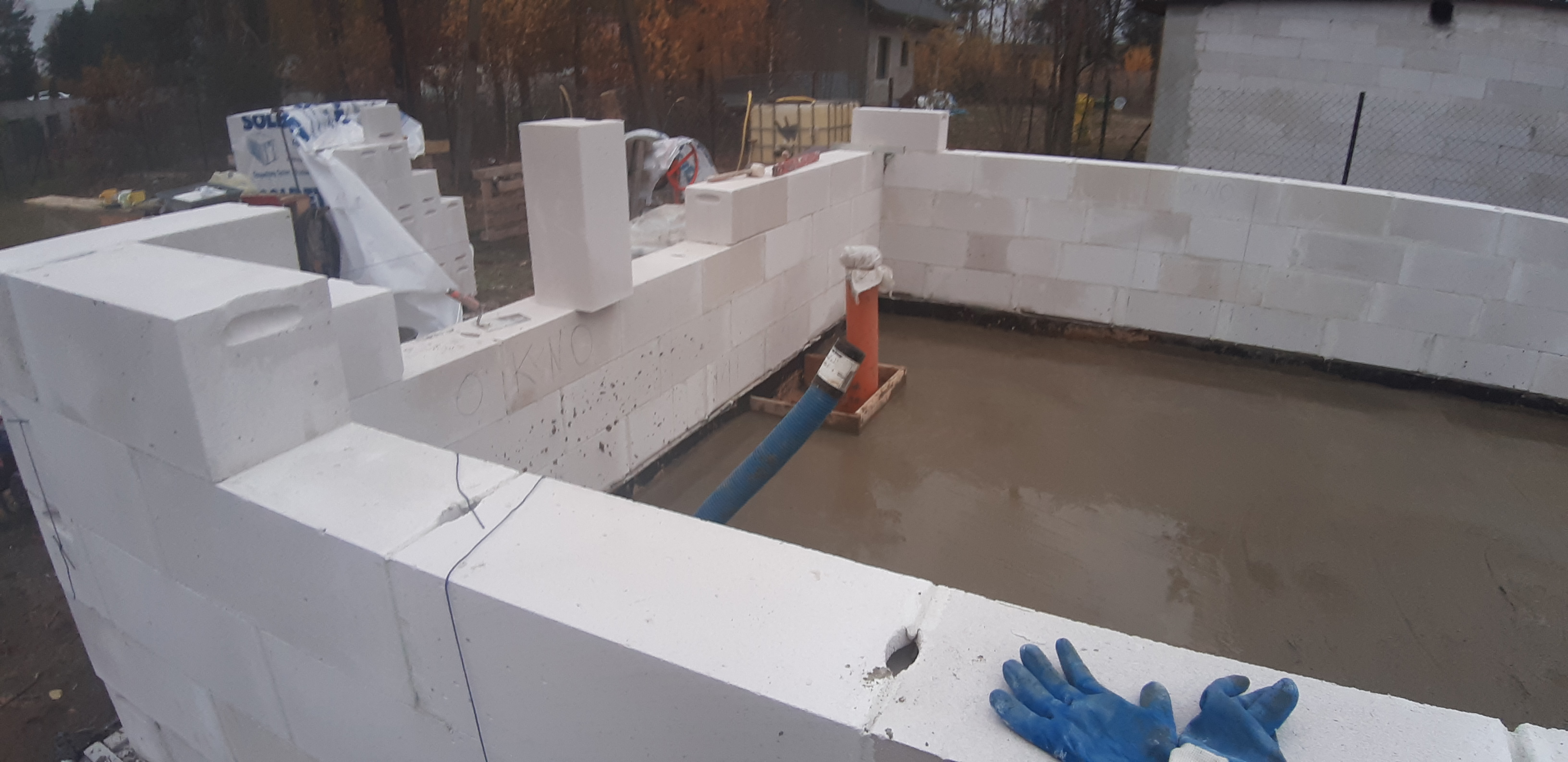 Wznoszone ściany fundamentowe z bloczków betonowych, wewnątrz wylany beton, widoczne niebieskie rękawice robocze i węże do pompowania betonu.