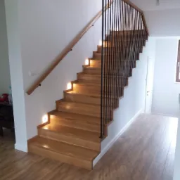 Drewniane schody z podświetleniem stopni i czarną, pionową balustradą w nowoczesnym domu.