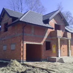 Niedokończony budynek jednorodzinny z czerwonej cegły, z widoczną konstrukcją dachu pokrytego szarą dachówką, otwartym garażem i schodami wejściowymi, na placu budowy z kopcami ziemi.