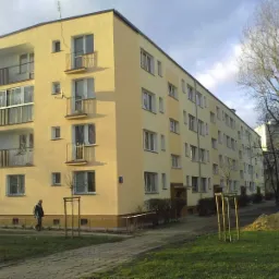 Żółty blok mieszkalny z balkonami, widoczny mężczyzna z psem na trawniku, drzewa bez liści i sterta piasku.