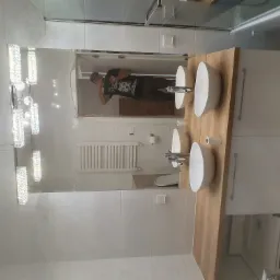 Nowoczesna łazienka z podwójną umywalką nablatową, drewnianym blatem i kryształowym oświetleniem sufitowym, widok z perspektywy lustra, w którym odbija się osoba robiąca zdjęcie oraz fragment...