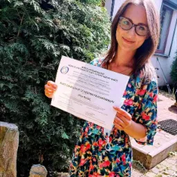 Uśmiechnięta kobieta we Wrocławiu prezentuje certyfikat 'Basic Consultant of Positive Psychotherapy' wydany przez World Association for Positive and Transcultural Psychotherapy (WAPP). W tle...