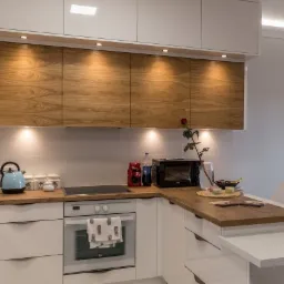 Nowoczesna kuchnia z białymi szafkami dolnymi i górnymi z drewnianymi frontami, oświetleniem punktowym, drewnianym blatem i wyspą z białym blatem, na blacie czajnik, mikrofalówka, napoje i wazon...