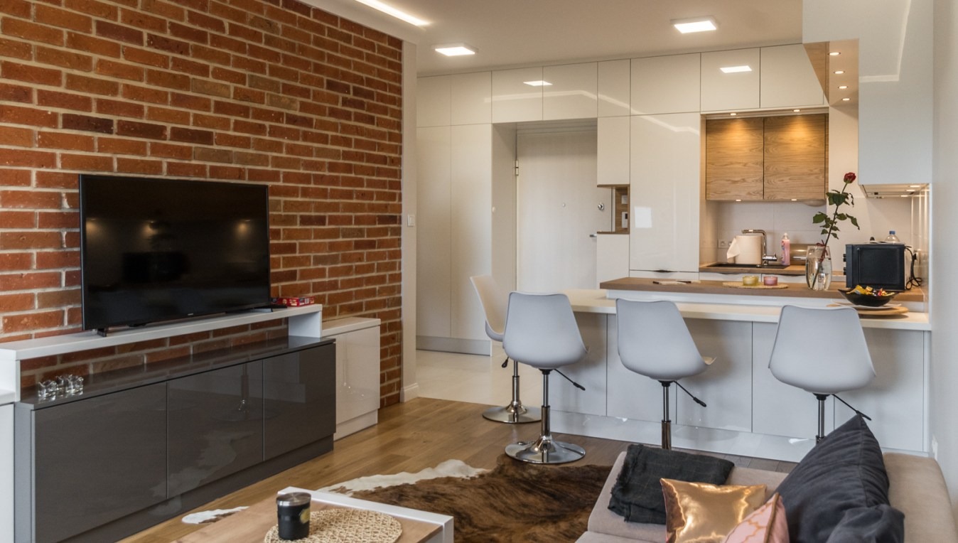 Nowoczesne wnętrze mieszkania z ceglaną ścianą, szarymi szafkami RTV, drewnianą podłogą i aneksem kuchennym z barkiem oraz szarymi krzesłami barowymi.