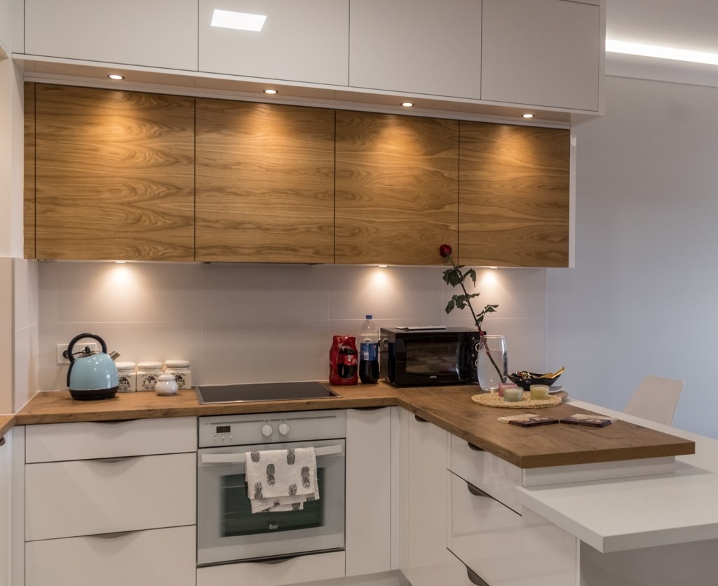 Nowoczesna kuchnia z białymi szafkami dolnymi i górnymi z drewnianymi frontami, oświetleniem punktowym, drewnianym blatem i wyspą z białym blatem, na blacie czajnik, mikrofalówka, napoje i wazon...