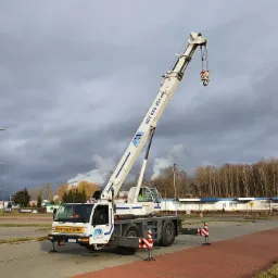 Żuraw samochodowy z wysuniętym ramieniem na tle pochmurnego nieba i zabudowań przemysłowych, z widocznym napisem 'Convoi Exceptionnel' na kabinie.