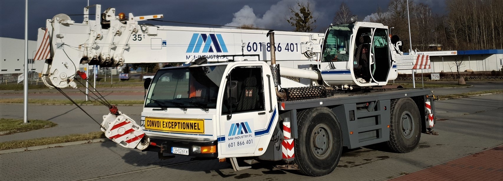 Biały dźwig samochodowy z otwartymi drzwiami kabiny, zaparkowany na placu z widocznym znakiem 'Convoi Exceptionnel' i logo firmy MM-Industry.