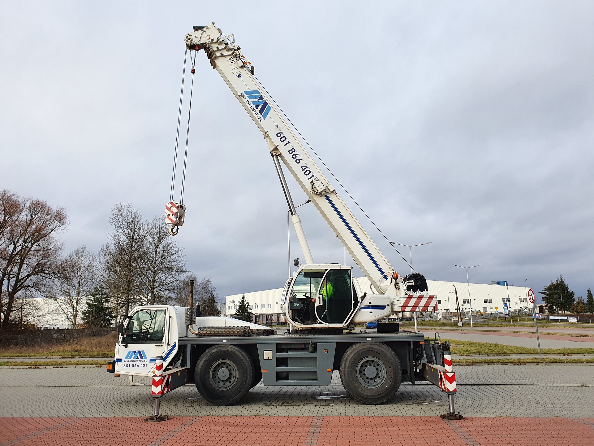Mobilny dźwig samochodowy z wysięgnikiem teleskopowym, zaparkowany na kostce brukowej, z widocznym operatorem w kabinie i budynkiem przemysłowym w tle.